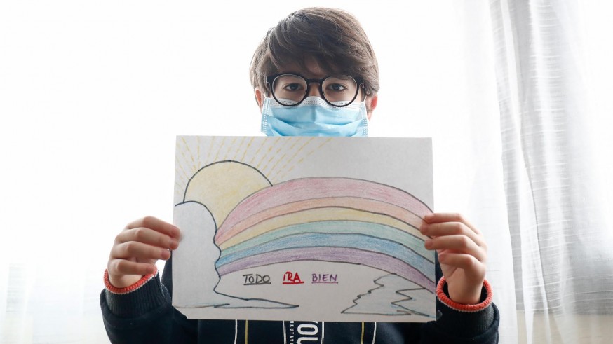 Un niño con mascarilla enseña un dibujo que ha hecho de un arcoiris y la frase 'Todo irá bien'